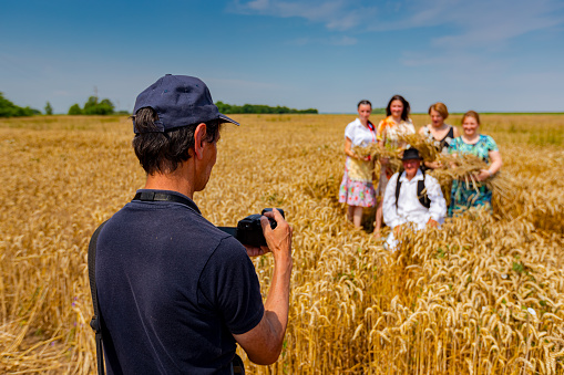 Muzlja, Vojvodina, Serbia, - July 01, 2023; Photographer is holding photo camera, he taking pictures, looking at camera on family in white folk costume as standing in field of wheat