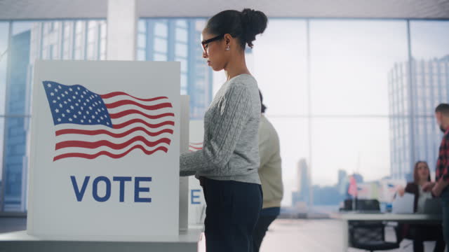 Young African American Woman Filling Out a Ballot Next to a Voting Booth on the Day of Elections in the United States. Diverse Men and Women Voting for Presidential Candidates in a Polling Station
