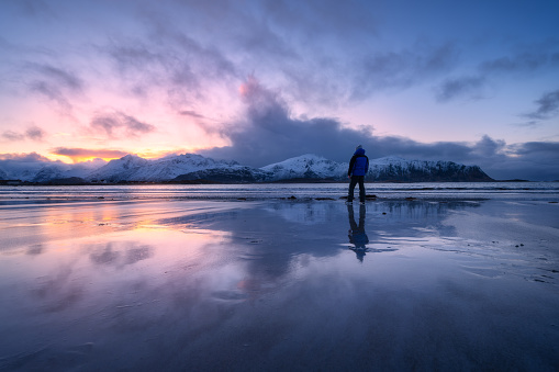Man on the sandy beach against snowy mountains and colorful sky at sunset in winter. Lofoten islands, Norway. Landscape with silhouette of a guy, sea, reflection in water, purple sky with pink clouds