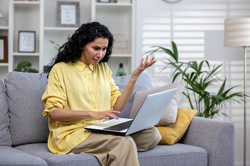 Curly female Hindu student sitting at home on sofa with laptop on lap, unhappy with non-working computer, poor internet connection. Attractive woman having problems working from home.