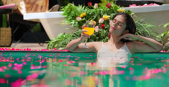 Indian beautiful woman standing in swimming pool edge hold glass drinking juice at outdoor home. Happy female bathing in luxury resort hotel doing relaxation pose in blue water enjoy summer vacation