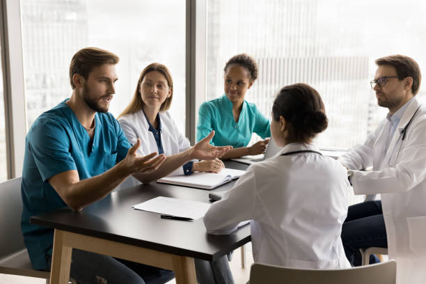 Confident young surgeon man in uniform talking to multiethnic colleagues Confident young surgeon man in blue medic uniform talking to multiethnic colleagues in hospital meeting room, speaking to listening diverse doctors at table, offering ideas for clinic teamwork medical occupation stock pictures, royalty-free photos & images