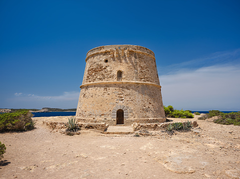 View of Torre d'en Rovira, an ancient defence tower located on the western coast of Ibiza, not far from Platges de Comte and Cala Bassa, built during the 18th century to watch over the bay of Sant Antoni de Portmany (also known as San Antonio Abad). The dazzling bright light of a Mediterranean summer noon, an intense blue sky, picturesque clouds, deep blue waters as far as the eye can see, lush bushes of Mediterranean scrub, agave plants. High level of detail, natural rendition, realistic feel. Developed from RAW.