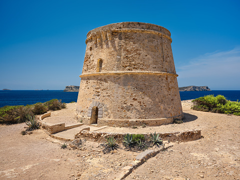 View of Torre d'en Rovira, an ancient defence tower located on the western coast of Ibiza, not far from Platges de Comte and Cala Bassa, built during the 18th century to watch over the bay of Sant Antoni de Portmany (also known as San Antonio Abad). The dazzling bright light of a Mediterranean summer noon, an intense blue sky, picturesque clouds, deep blue waters as far as the eye can see, lush bushes of Mediterranean scrub, agave plants. High level of detail, natural rendition, realistic feel. Developed from RAW.