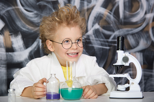 Happy child with glasses holds samples of chemical liquid in a flask with laboratory utensils on a chalkboard background. Child in a scientist's robe has fun doing experiments in the laboratory