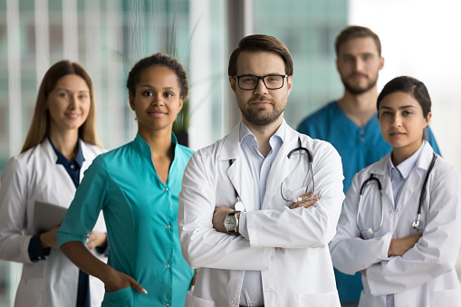 Confident head doctor wearing white coat and glasses standing in front of diverse team of medical colleagues, keeping arms crossed, looking at camera. Multiethnic professionals posing for portrait