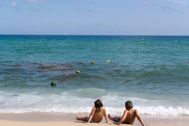 image in full sun of two children sitting on the sand where the waves of the blue sea break while looking at the horizon