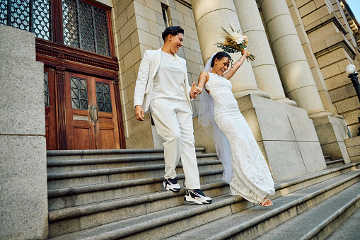A radiant couple in wedding attire laugh and hold hands against the grand columns of a city building, their joy palpable in the golden sunlight.