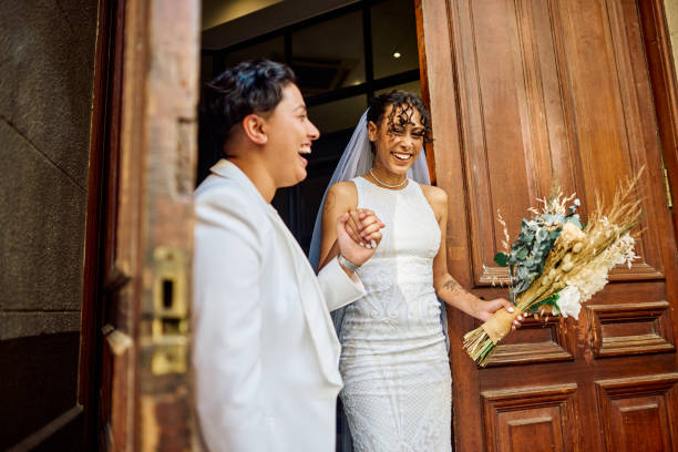LGBTQIA couple celebrating their love with an elopement wedding in the city. A radiant couple in wedding attire laugh and hold hands exiting a city building, their joy palpable in the golden sunlight. bride holding bouquet stock pictures, royalty-free photos & images