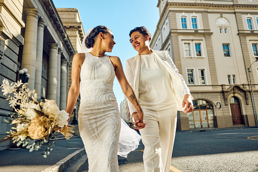 Two brides in elegant wedding garments holding hands and looking into each other's eyes with affection on a sunlit city sidewalk, their love glowing as bright as the day.
