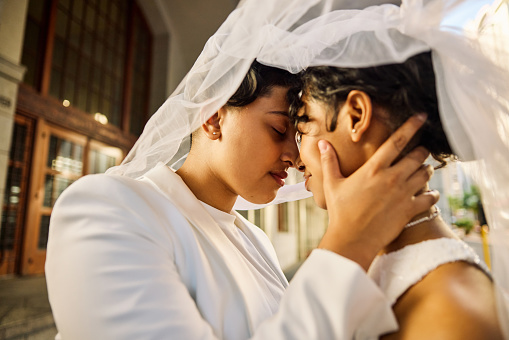 In a quiet, sunlit moment, two brides lean in close, their foreheads touching gently, as they share a serene and intimate embrace before the bustling excitement of their wedding day.