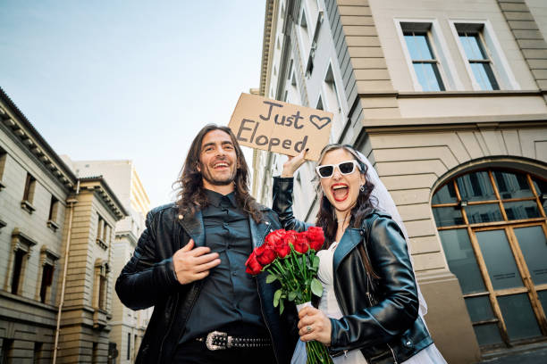 Overjoyed couple celebrating their elopement wedding in the city. A cheerful couple showcases their spontaneous spirit in the city, the bride holding a bouquet of red roses, as they proudly display their 'Just Eloped' sign, their smiles as bright as the day. eloping stock pictures, royalty-free photos & images