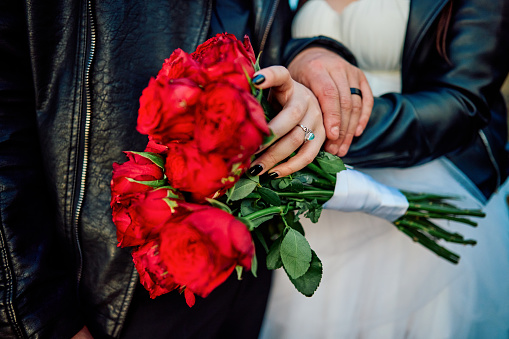 A close-up view of a couple's hands gently clasped, showcasing their wedding rings and a bouquet of deep red roses, embodying the romantic essence of their special day.