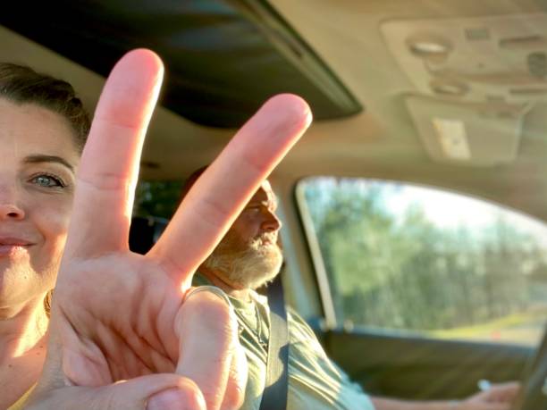 Married couple in a car interior, having fun on a road trip. The man, who is belted in and driving is a husky bearded mid age man, he is slightly defocused in the background, with the woman’s face, also mid age, wearing a smile and her hands put up in a peace sign, being the main focus. Both are having fun and riding in the car together.