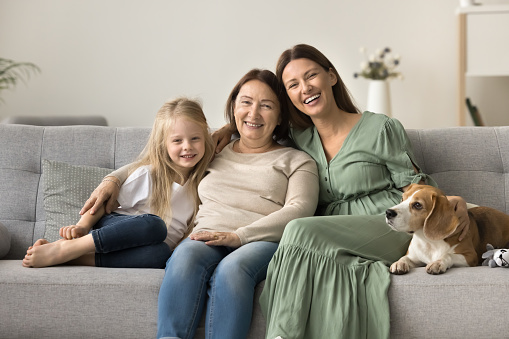 Cheerful pretty mom, grandma, happy preschool girl and dog resting on soft couch together, stroking pet, looking at camera for family portrait, smiling, laughing, enjoying relations, affection