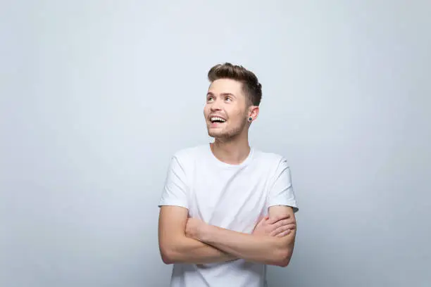 Surprised young man wearing white t-shirt standing with arms crossed and looking away. Studio shot, grey background.