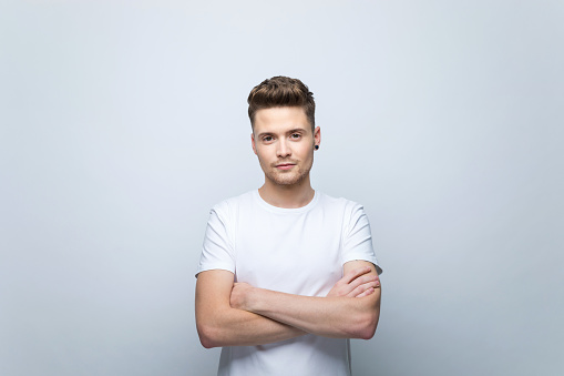 Friendly young man wearing white t-shirt standing with arms crossed and looking at camera. Studio shot, grey background.