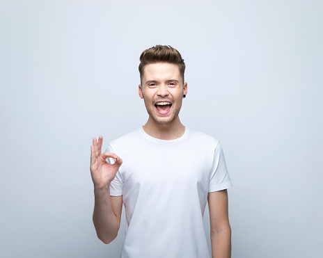 Happy young man wearing white t-shirt smiling at camera and showing ok sigh. Studio shot, grey background.