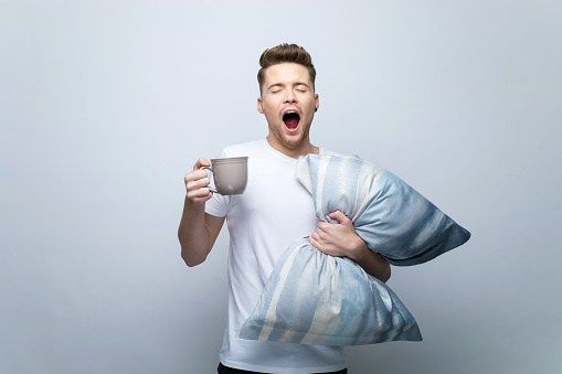 Young man wearing white t-shirt yawning with eyes closed, holding pillow and mug with coffee. Studio shot, grey background.