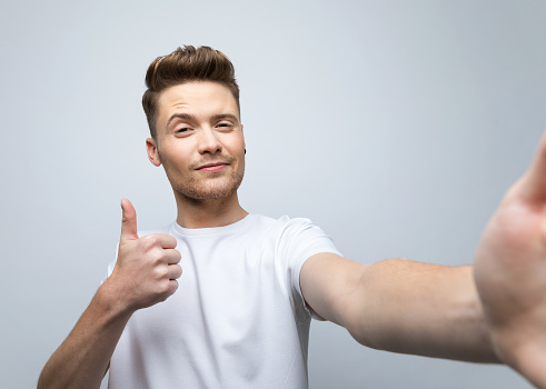 Happy young man wearing white t-shirt smiling at camera with thumb up, taking selfie. Studio shot, grey background.
