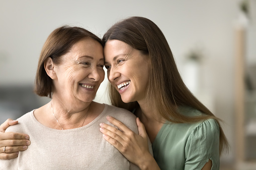 Happy young adult daughter woman hugging elder mature mom from behind, touching mothers shoulders, talking, laughing with head touches, enjoying family bonding, parent and child relationship