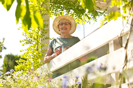 Outdoor portrait of smiling senior woman wearing straw hat with coffee cup on porch, looking at camera.