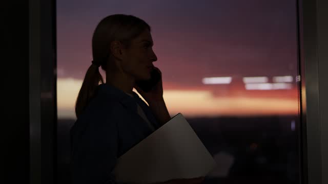 Close up of businesswoman standing and talking on smartphone while holding laptop by window with great sunset view. Female boss discussing working moments with client in the evening in modern house.