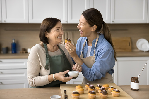 Joyful senior mom and adult daughter having fun while cooking dessert, decorating chocolate muffins, tasting cupcakes. Happy young woman giving sweet cupcake to mother for biting
