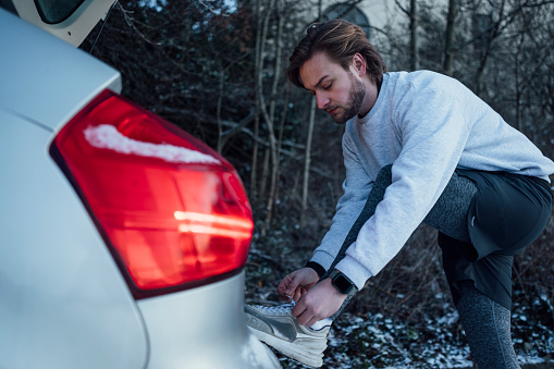 Side angle view of a young male adult standing at the back of his car. He is prepping for a run and tying his shoelaces.