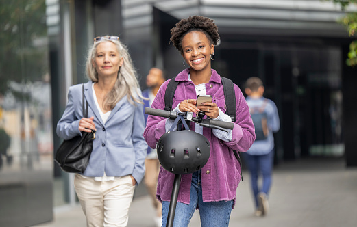 Portrait of smiling woman with helmet, holding smartphone and with e-scooter. Modern lifestyle concept.