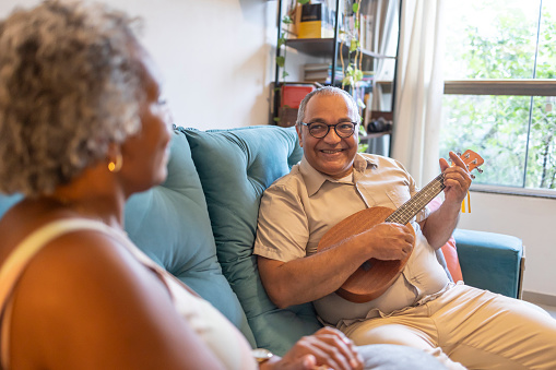 middle-aged latin man plays ukulele for black woman in living room