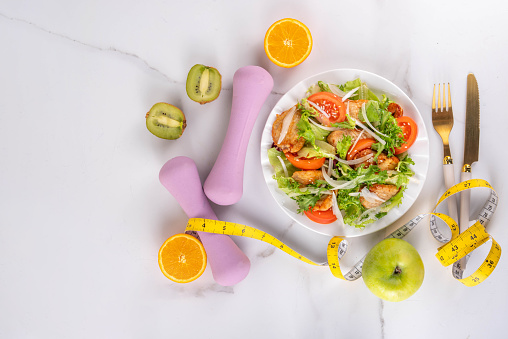 Weight loss, Healthy lifestyle, diet food, sport and fitness concept. Fresh balanced vegetable and chicken breast salad with fruit, fitness equipment, dumbbell, measuring tape on a white background