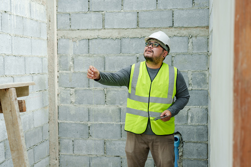 Asian male construction engineer, wearing full clothing, safety helmet, reflective vest, holding laptop in hand, checking work site for completeness, following construction plans.