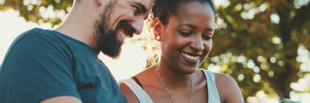 istock Happy interracial couple talking while sitting on bench, using cellphone, Panorama 1970247960