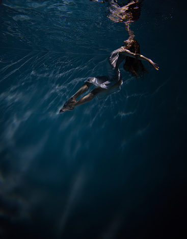 Underwater shoot of beautiful ballerina in white dress swimming and dancing in water through sunbeams. Ballerina against water background.
