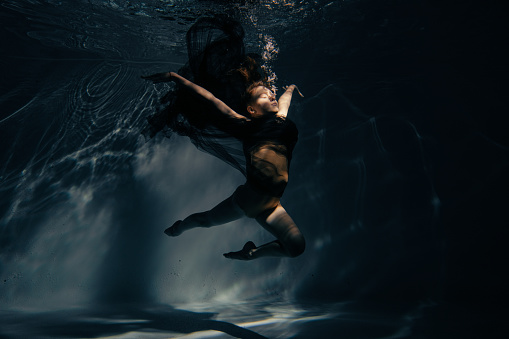 Underwater shoot of beautiful ballerina in black dress swimming and dancing in water through sunbeams. Ballerina against water background.