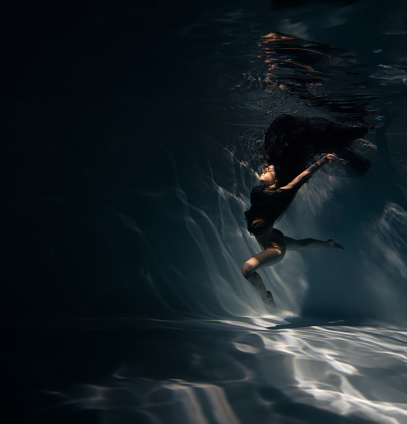 Underwater shoot of beautiful ballerina in black dress swimming and dancing in water through sunbeams. Ballerina against water background.