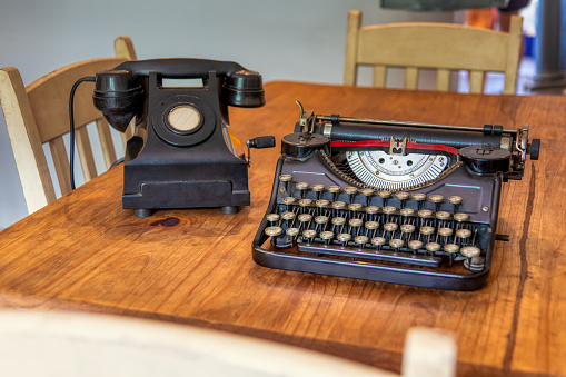 old black typewriter on a vintage wooden table at home , aged rotary crank telephone in the background