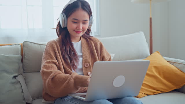 Attractive happy young asian woman wearing headphones looking at a laptop screen video call learning online on internet, listening online teacher or chatting with her friend.
