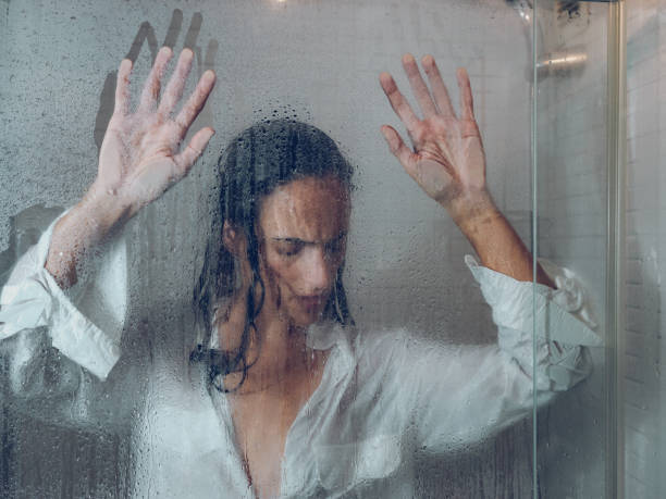 Unhappy woman near misted glass door in shower Through glass of sad female with black hair in wet wear touching transparent door of shower cabin in light bathroom fog transparent stock pictures, royalty-free photos & images