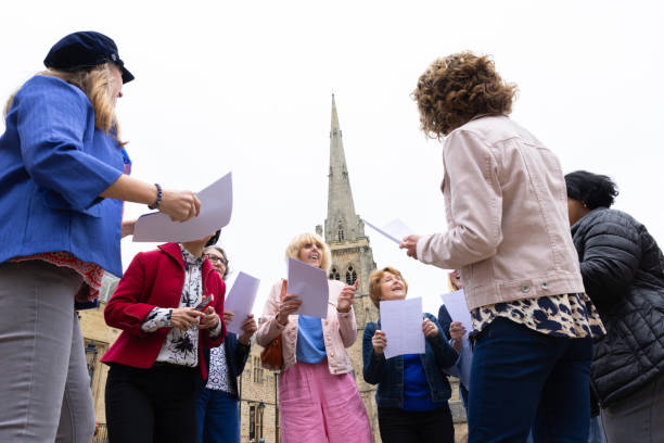 A low-angle three-quarter length handheld shot of a group of senior women singing in a group choir with each other wearing casual clothing. They are outside located Durham, England.