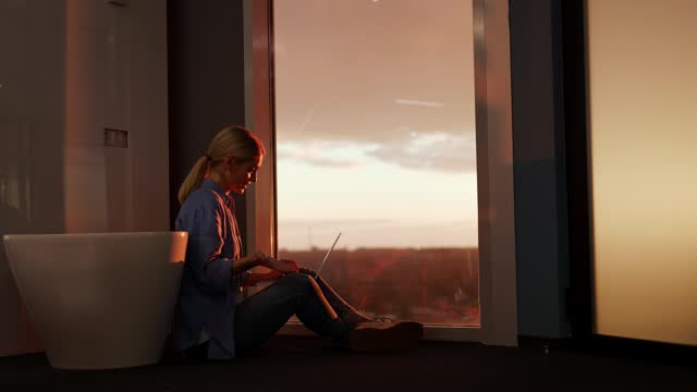 Woman having online conference on laptop while sitting on floor by panoramic window with stunning view. Businesswoman using portable computer for meeting with clients and discussing work in evening.