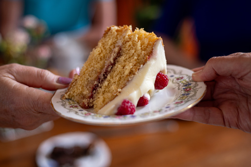 Unrecognisable mature adults passing a plate with a piece of cake on it. The cake is sponge cake decorated with cream icing and raspberries. The rustic cafe is located in Durham, England.