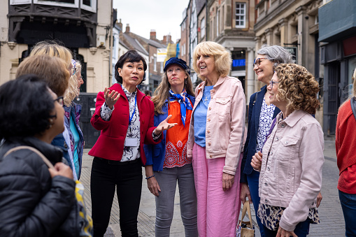 An over-the-shoulder three-quarter length shot of a group of women standing and listening with a tour guide in a historic English town. They are all wearing casual clothing located in Durham, England.