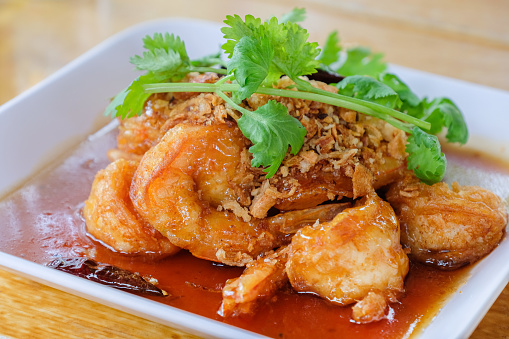 Crispy Prawn in Tamarind Glaze with fried garlic and coriander decorated in ceramic white plate served on bright wooden table background, Thai food
