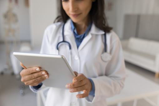 Cropped shot of young practitioner woman using digital tablet for communication, online help, working with professional application. Close up of gadget with doctor in blurred background