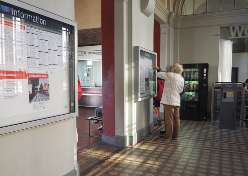 Ruedesheim, Germany – January 01, 2023: Old couple looking for departure times on information plan inside station building with historical floor tiles, man pointing with finger at plan
