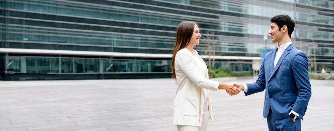 Two business associates shake hands in front of an office buildings, symbolizing a successful agreement or partnership initiative. Businessman and businesswoman made a deal, wide view, banner