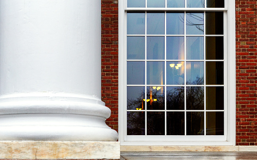 A close-up of a university's old classic building exterior featuring a white architectural column and a window through which is seen a dimly lit interior.
