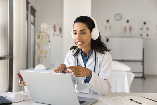 Young Latin doctor woman in wireless headphones speaking on video conference call at laptop, talking to patient online in hospital office, giving lecture, webinar to medical students